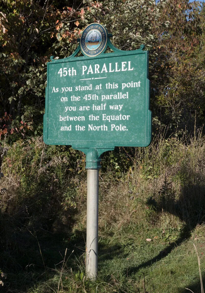 A colored photograph of a tall green sign with white lettering. The sign stands in a small clearing between green trees and bushes.