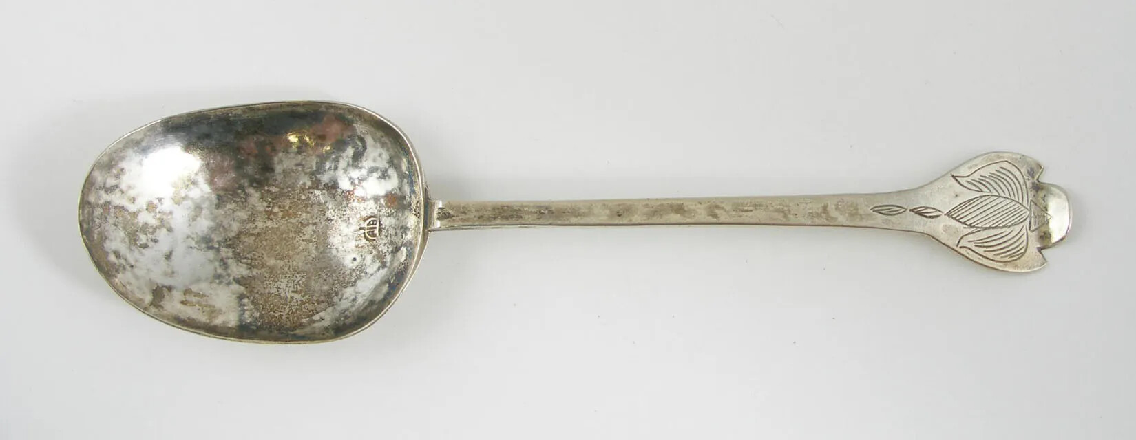 A silver-colored spoon, with a thin handle. On the end of the handle is a carving of three leaves.