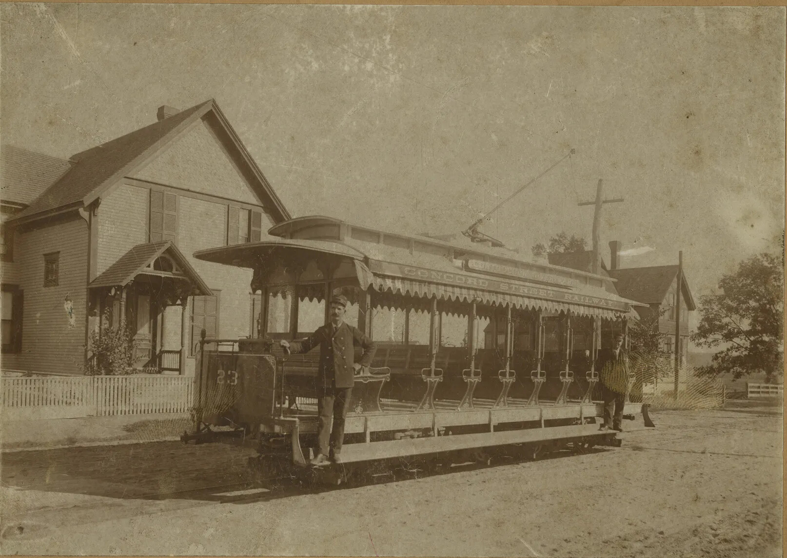 A black and white photograph of a passenger car sitting on a set of railroad tracks in front of a row of houses. The car has a roof, which is attached to a suspended wire by a tall metal stick. The sides of the car are opened, and the inside of the car is lined with benches. Two men stand on either side of the car on a raised platform.