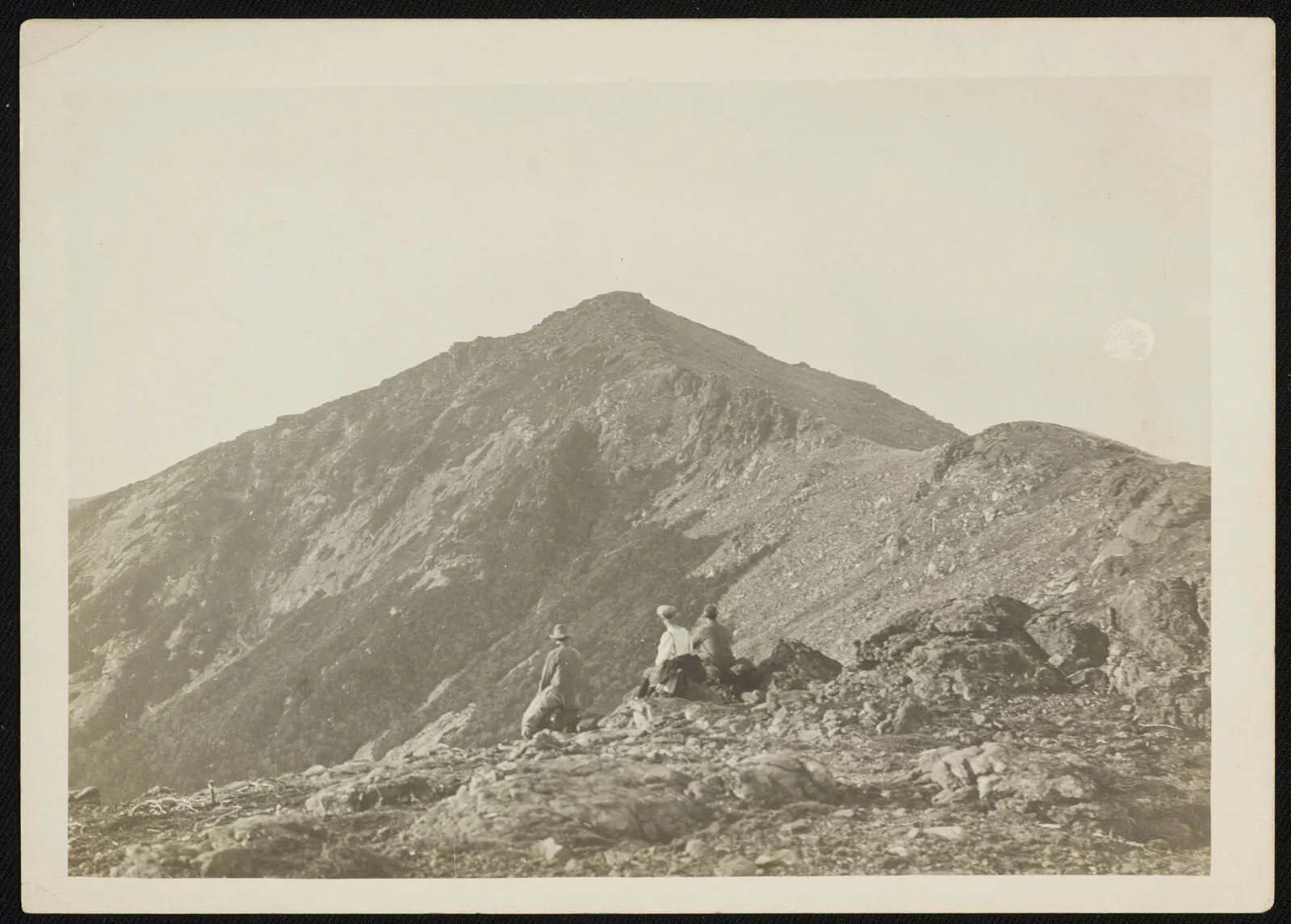 A black and white photograph of three people sitting on a rocky surface, facing a tall, pointed mountain.