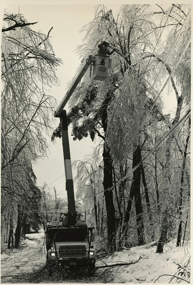 A black and white photograph of a truck parked along a snow-covered road, surrounded by snow and ice-covered trees. The truck has a large crane and lift. Inside the lift is a man bending near a broken tree branch.