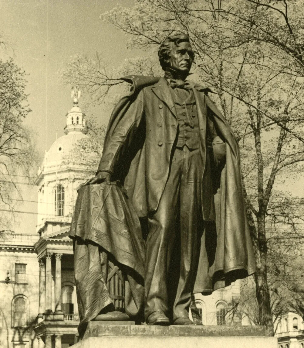 A tall statue of a man with his right hand on his right hip, and his left hand on a short pillar covered in an American flag. He wears a high-collared shirt, necktie, vest, pants, and long coat. Over his shoulders is draped a large collared coat.