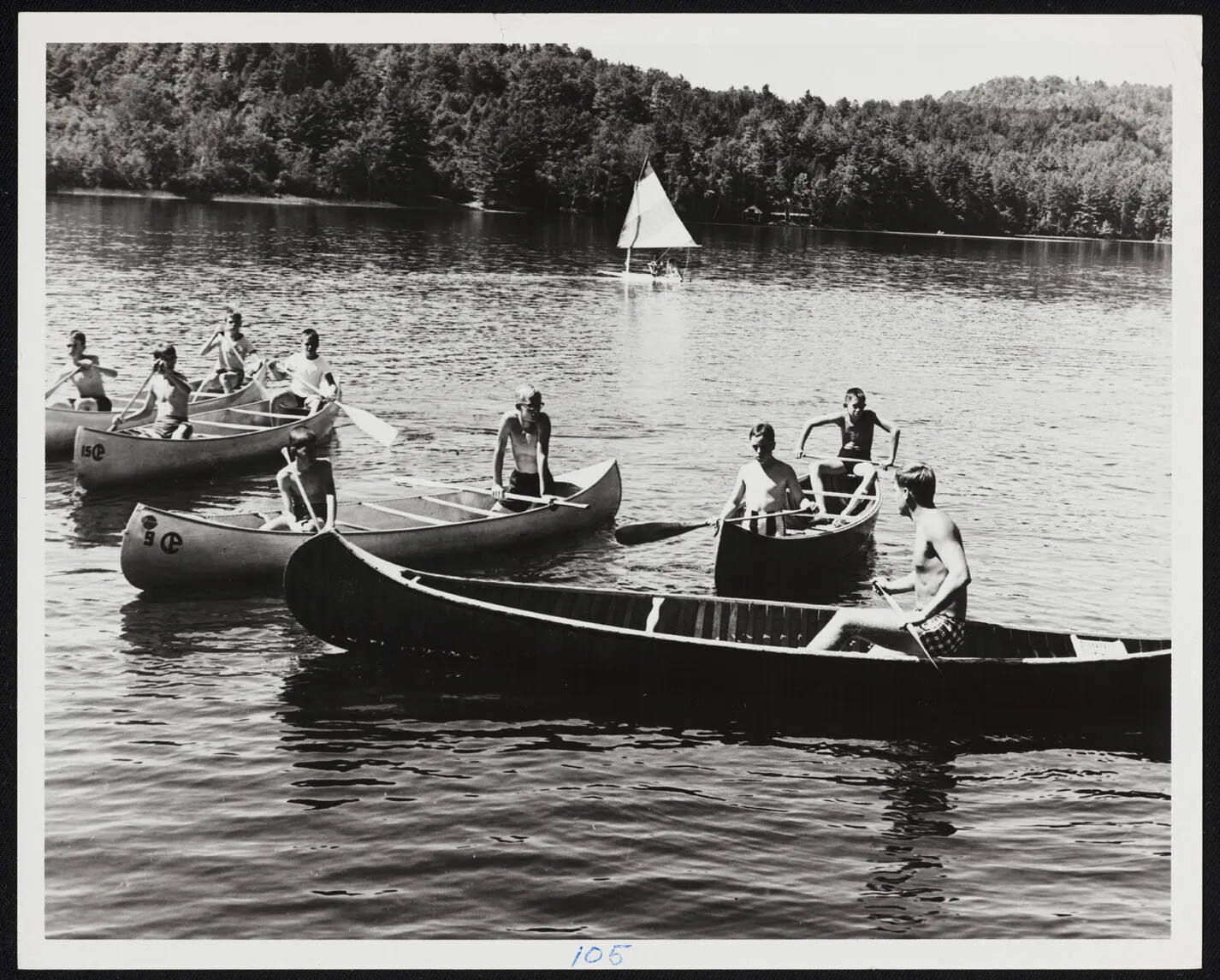 A black and white photograph of various groups of boys in large canoes on a body of water. There are mountains in the background and a small sailboat sails just behind the canoes.