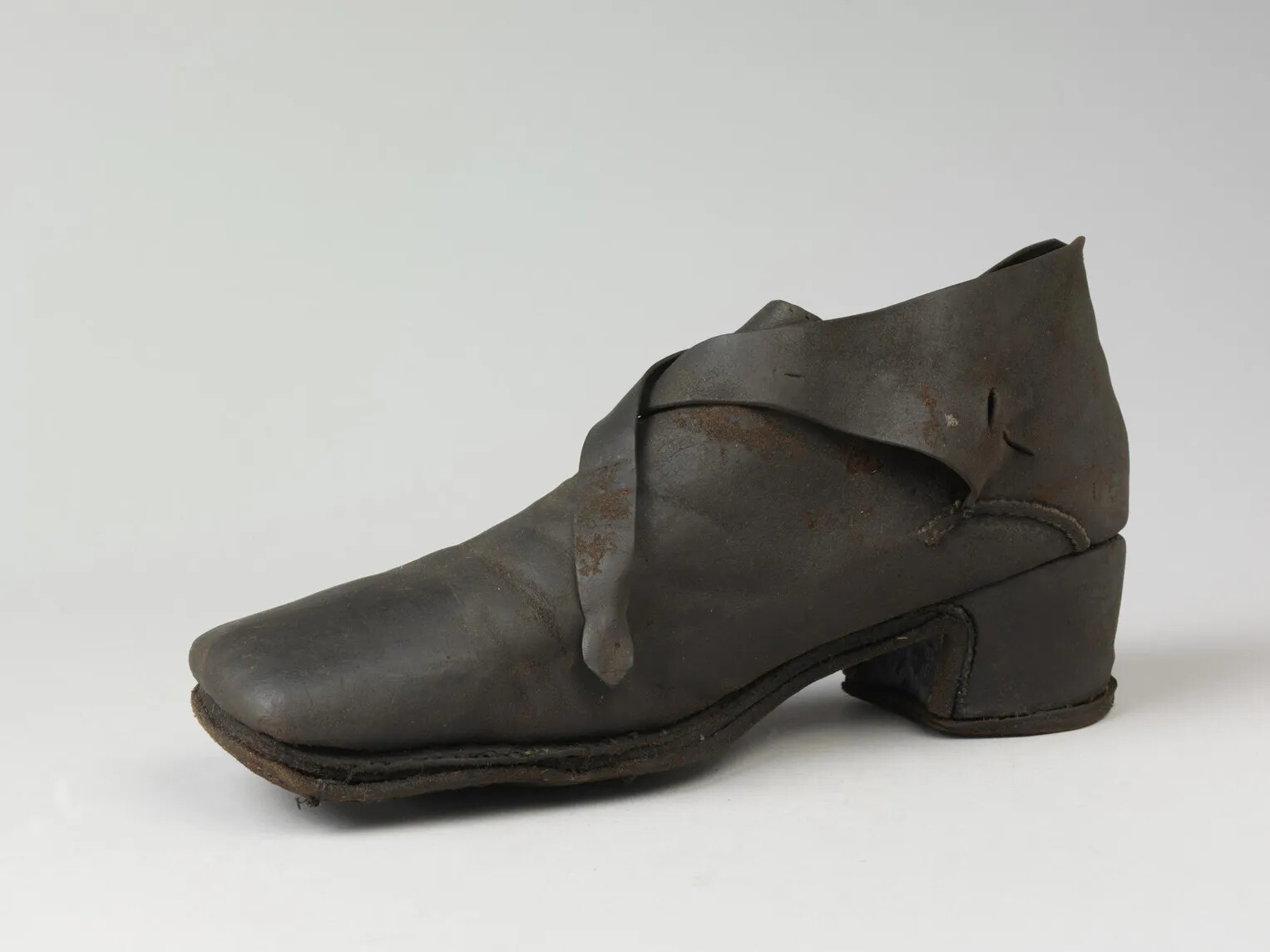 A brown leather shoe, with the toe pointed to the left and the heal pointed to the right. The shoe is fastened with two long leather straps, which cross each other along the top of the shoe.