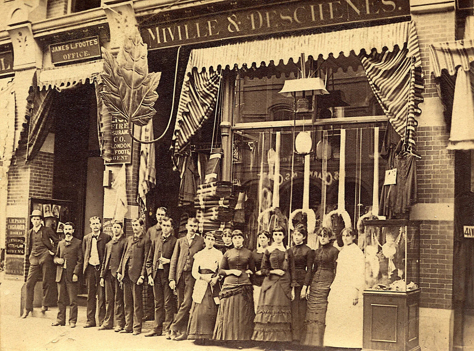 A black and white photograph of a group of men and women standing in front of a store. The men wear dark colored jackets and pants, and the women wear long dresses of various designs. Above the store is a sign that reads "MIVILLE & DESCHENES." A large maple leaf sign hangs out from the awning of the store into the street.