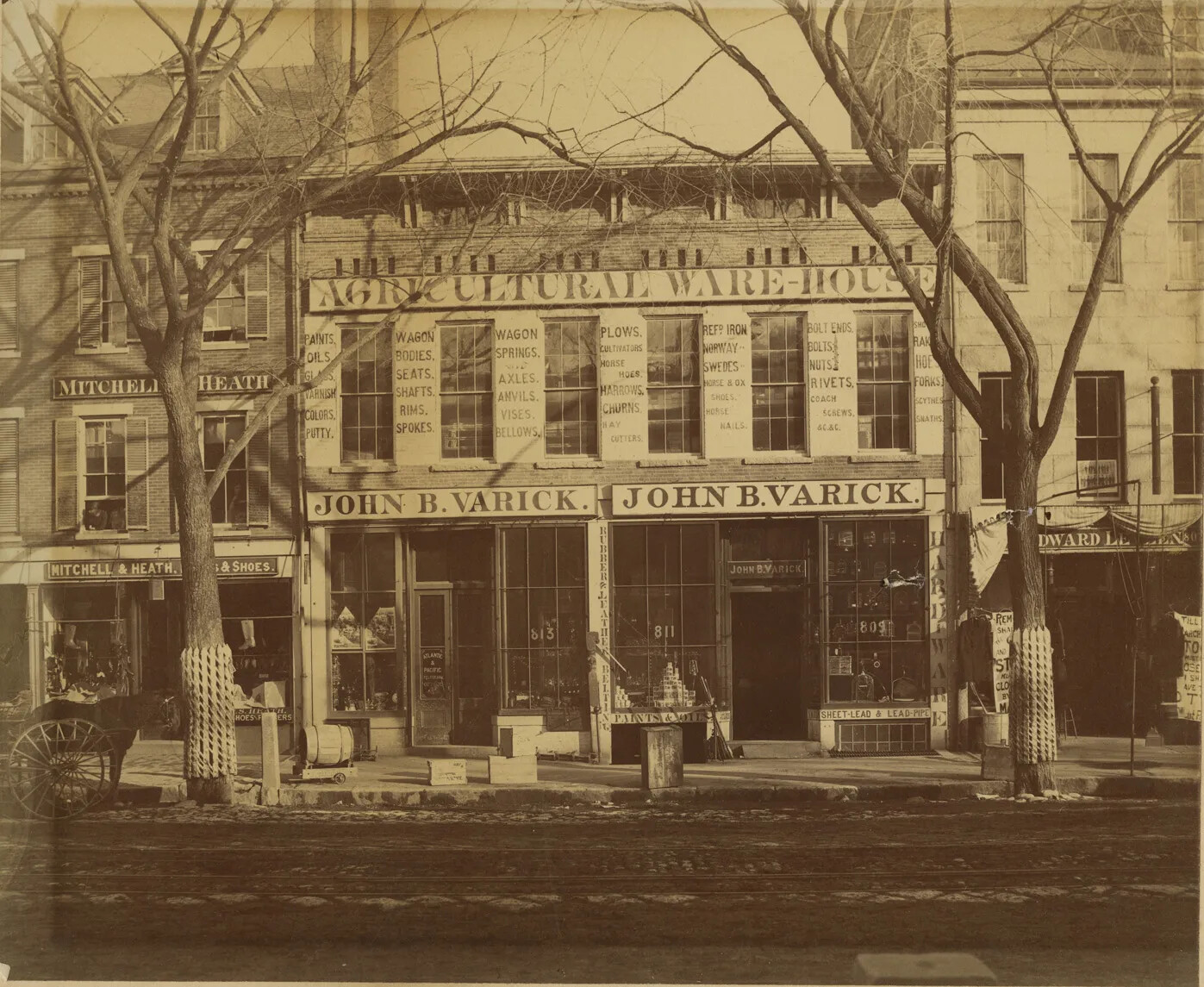 A black and white photograph of a two-storied store, in between two buildings of varying height on a street. Signs on the store read "AGRICULTURAL WAREHOUSE/JOHN B. VARICK."