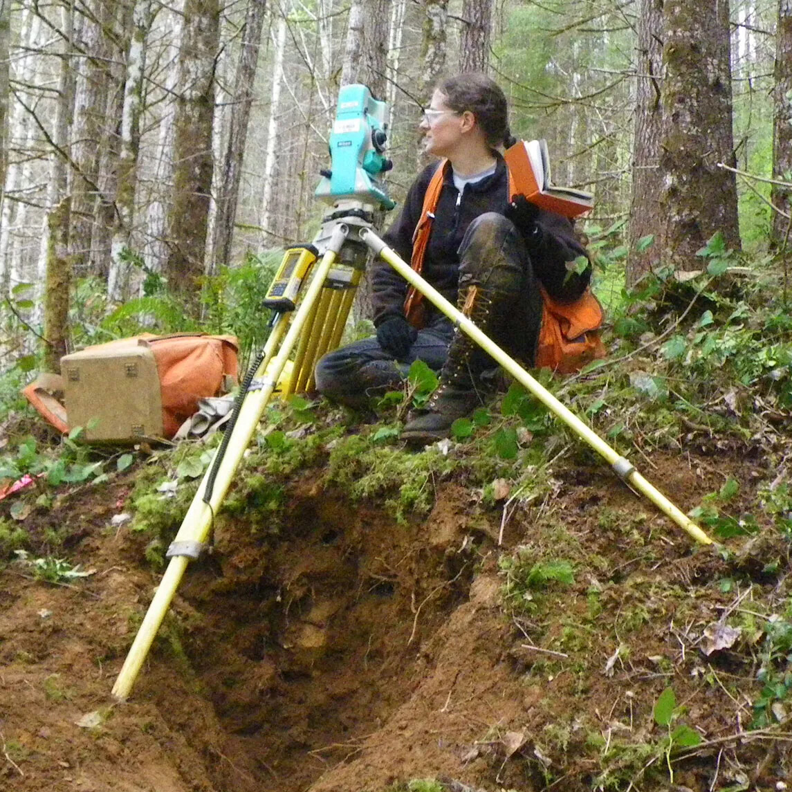 A colored photograph of a women wearing glasses and modern-day clothes sitting on a hill in a forest with a metal tripod. The women looks to the left and she holds an orange book in her right hand. To her left is a large orange backpack. 
