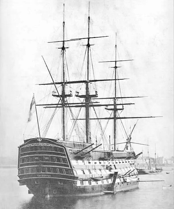 A black and white photograph of a large, multi-storied ship with three tall masts floating on a body of water.