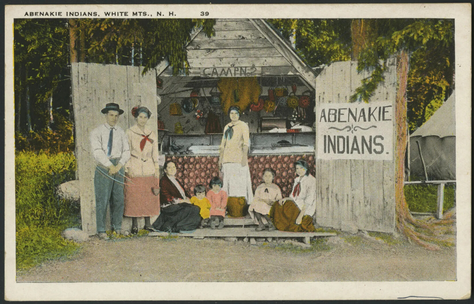 A colored drawing of a group of men, women, and children, who are standing and sitting in front of a small cabin. The cabin is filled with colorful bowls and other tools. A sign to the right of the cabin reads "ABENAKIE/INDIANS."