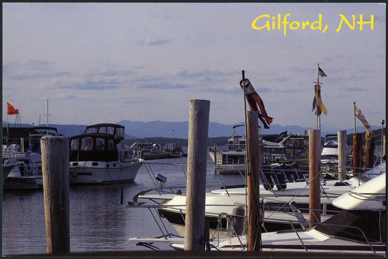 A colored horizontal postcard of a harbor with lots of boats parked at wooden docks. There are several circular flags placed on the tall dock posts. In the background are the outlines of mountains. The title of the postcard, placed in the upper right-hand corner, reads "Gilford, NH."