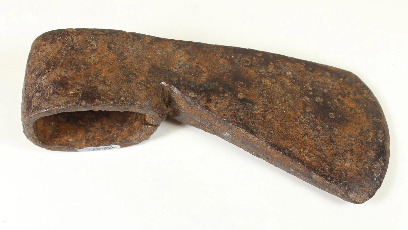 A rusty metal ax head, with a rounded edge on one side and a flattened edge on the other. The rounded side of the ax head is hollow, and has a hole where a handle would be placed.