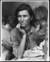 In a black and white photograph, a woman stares off into the distance, looking worried or tired. Her upper torso, shoulders, and head are seen, and her hand is holding her chin. Her hair is loosely pulled back, and she wears a blouse with cardigan. A small child is on either side of her with mussed hair and worn clothing. They have their heads on her shoulders and are facing away from the camera. 