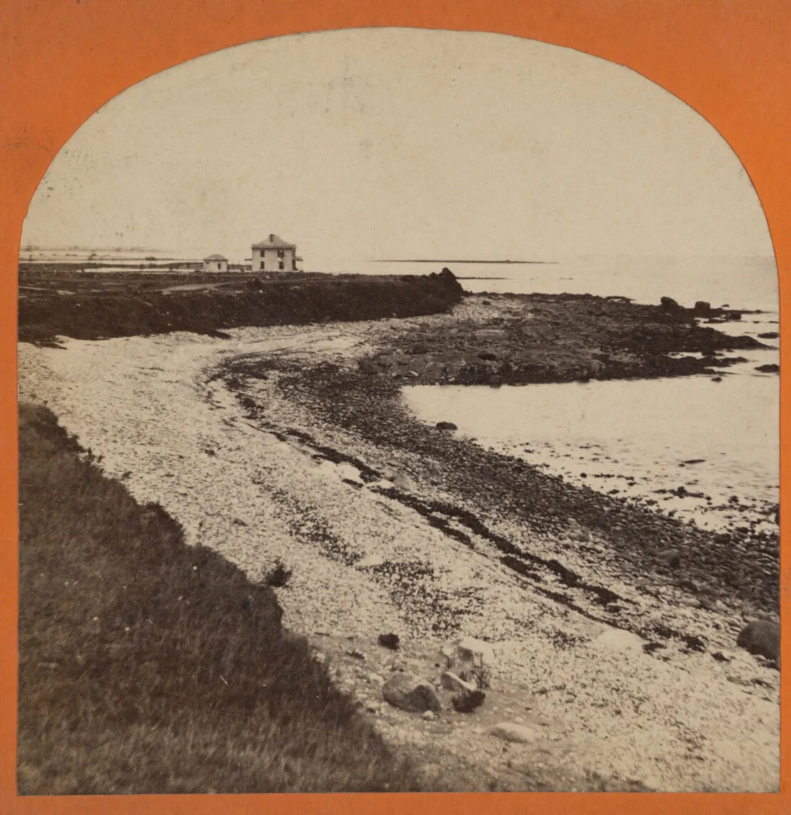 A black and white stereograph card with an image of a curved rocky beach. Above the beach is a raised grassy mound, and in the background, a two-storied house sits atop the raised mound.