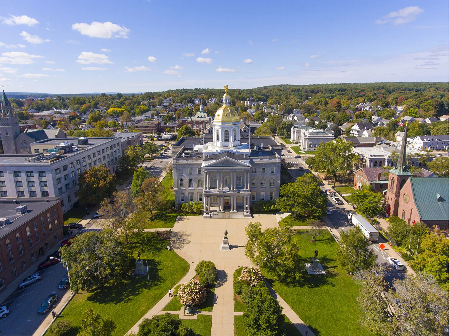 A colored photograph of a three-storied, domed building in the middle of a grassy lawn. Surrounding the lawn are multi-storied buildings. The building's dome is gold-colored and the rest of the building is gray. The front of the building juts out and is decorated with a row of columns.