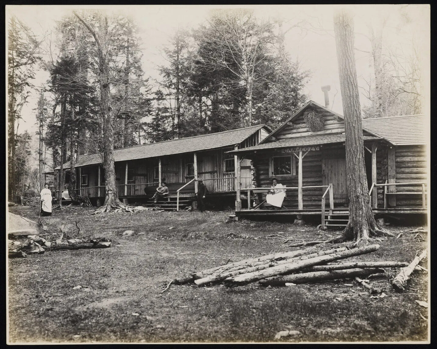 A black and white photograph of two log cabins with a forested back-drop. In front of each of the cabins is a single tree, and then a clearing with various logs scattered in piles around the yard. The cabins both have a covered front porch and a set of stairs leading up to the front door. The cabin to the left has a woman in a long dress off to its left, a man sitting on the stairs of the porch, and a man with a hat standing to the right of the stairs. The cabin in the right-hand side has a young woman in a light colored dress sitting on the porch, next to an empty chair. Above her is a sign, reading “CAMP COZY”.
