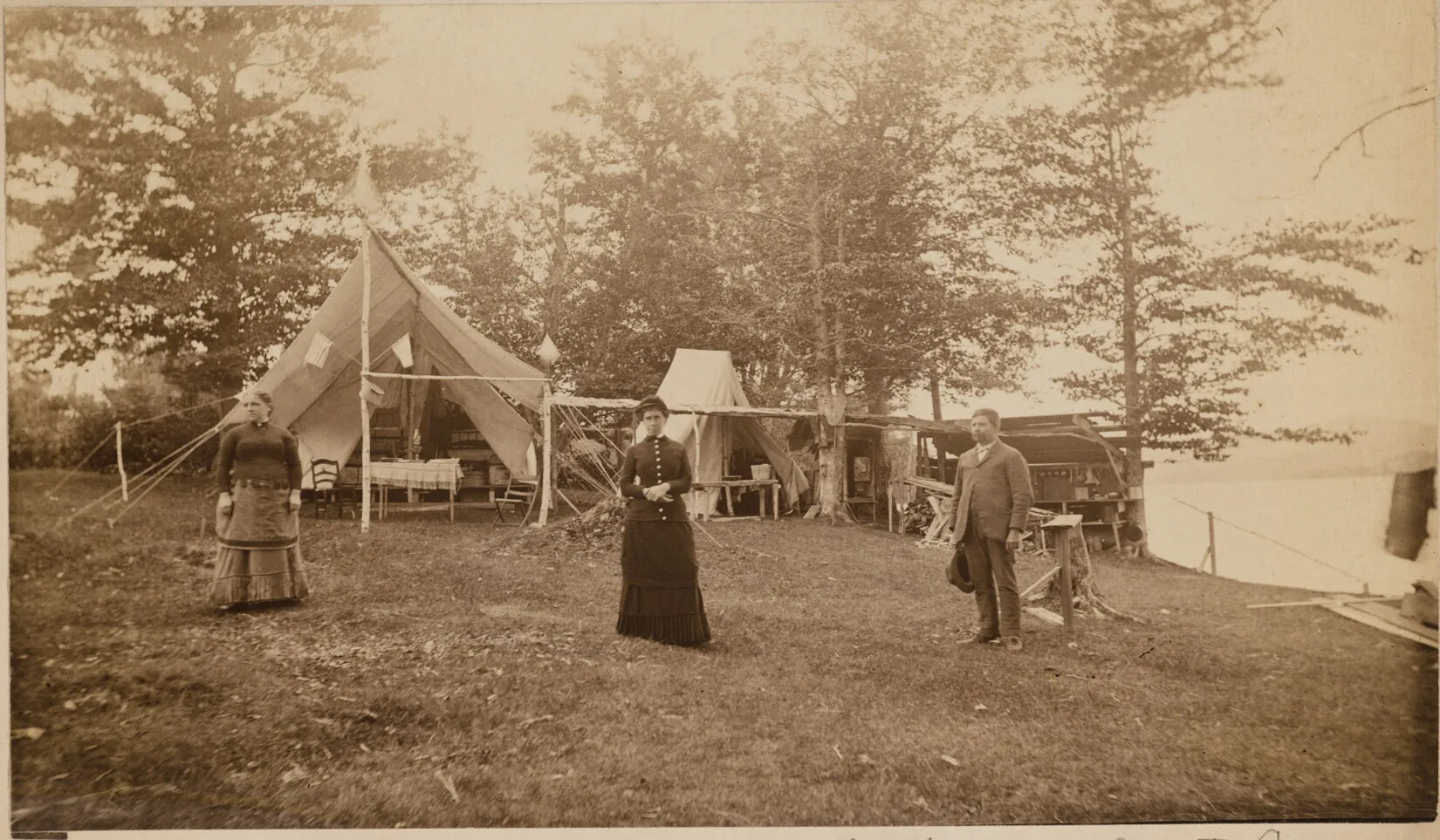 A black and white photograph of two women and one man standing in front of a camp site. The women are wearing long dresses and the man is wearing pants, a collared shirt, and a jacket, and holds his hat in his left hand. The camp site has two white tents. The larger tent, in the left-hand side of the photograph, has a dining room table with a tablecloth and has two chairs placed on either side of it. Towards the back of this tent, two beds can also be seen. The smaller tent, in the photograph’s center, is partially closed, and only a bench can be seen inside. Behind the two tents are tall trees, and in the right-hand corner, a body of water and a mountain are seen in the distance.