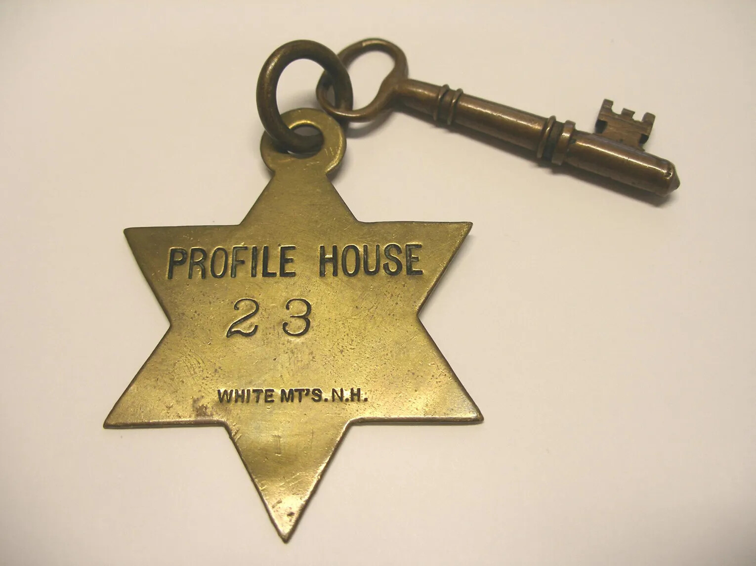 A brass colored key and a tag in the shape of a six-pointed star. The tag has three lines of indented text which reads: "PROFILE HOUSE/23/WHITE MT'S. N.H." On the back of the tag, there are two circles of curved, indented text. The text reads: "IF CARRIED AWAY RETURN/BY/MAIL/POSTAGE/2/CENTS/UNSEALED."