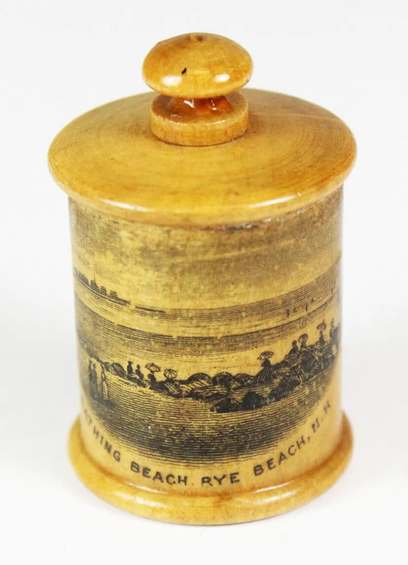 A wooden cylinder with a lid. There is an etching of people standing along the shore of a beach on the front of the cylinder.
