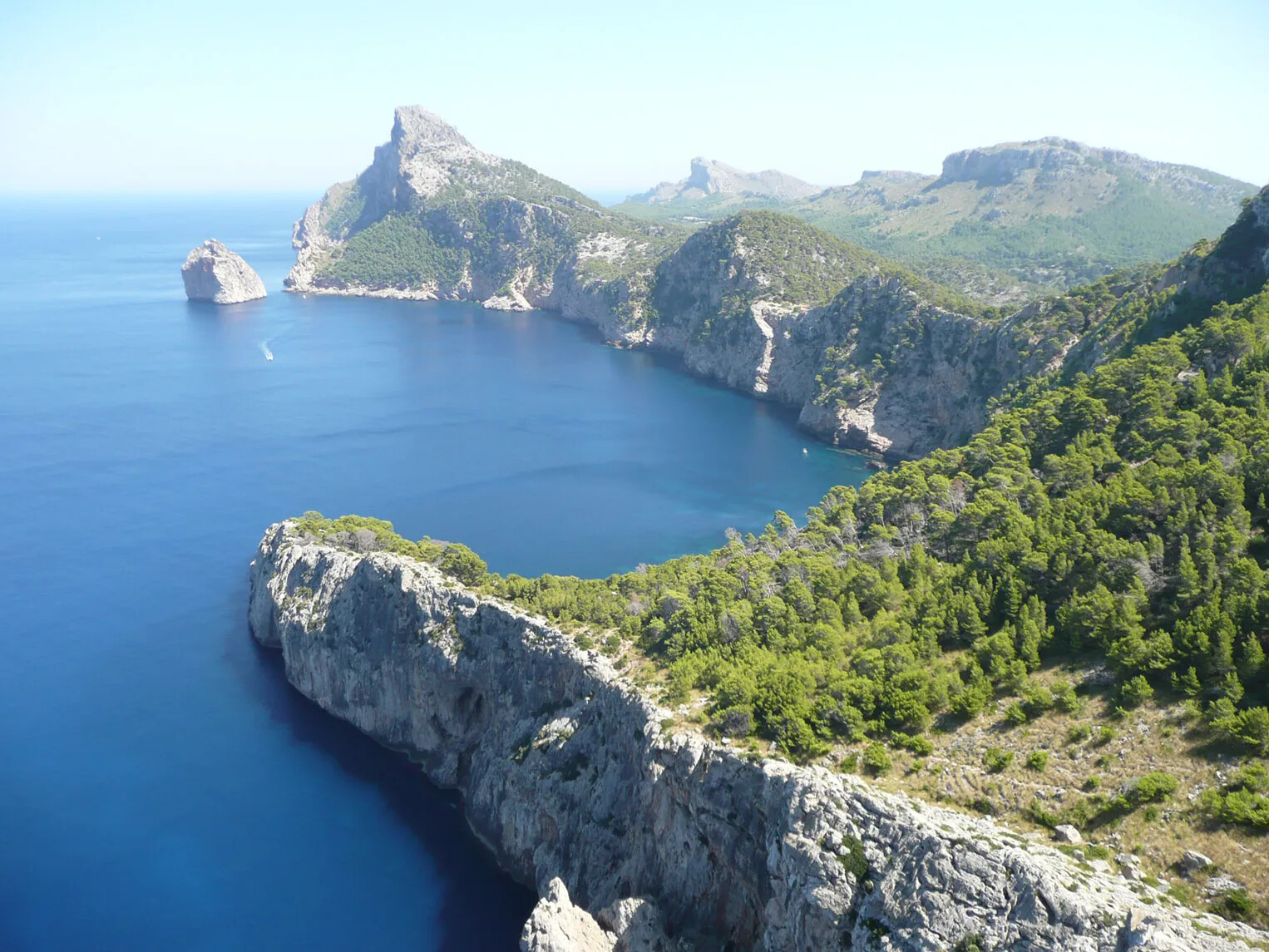 A colored photograph of tall mountain cliffs along a blue body of water. The mountains are gray and brown, and have green trees along the tops.
