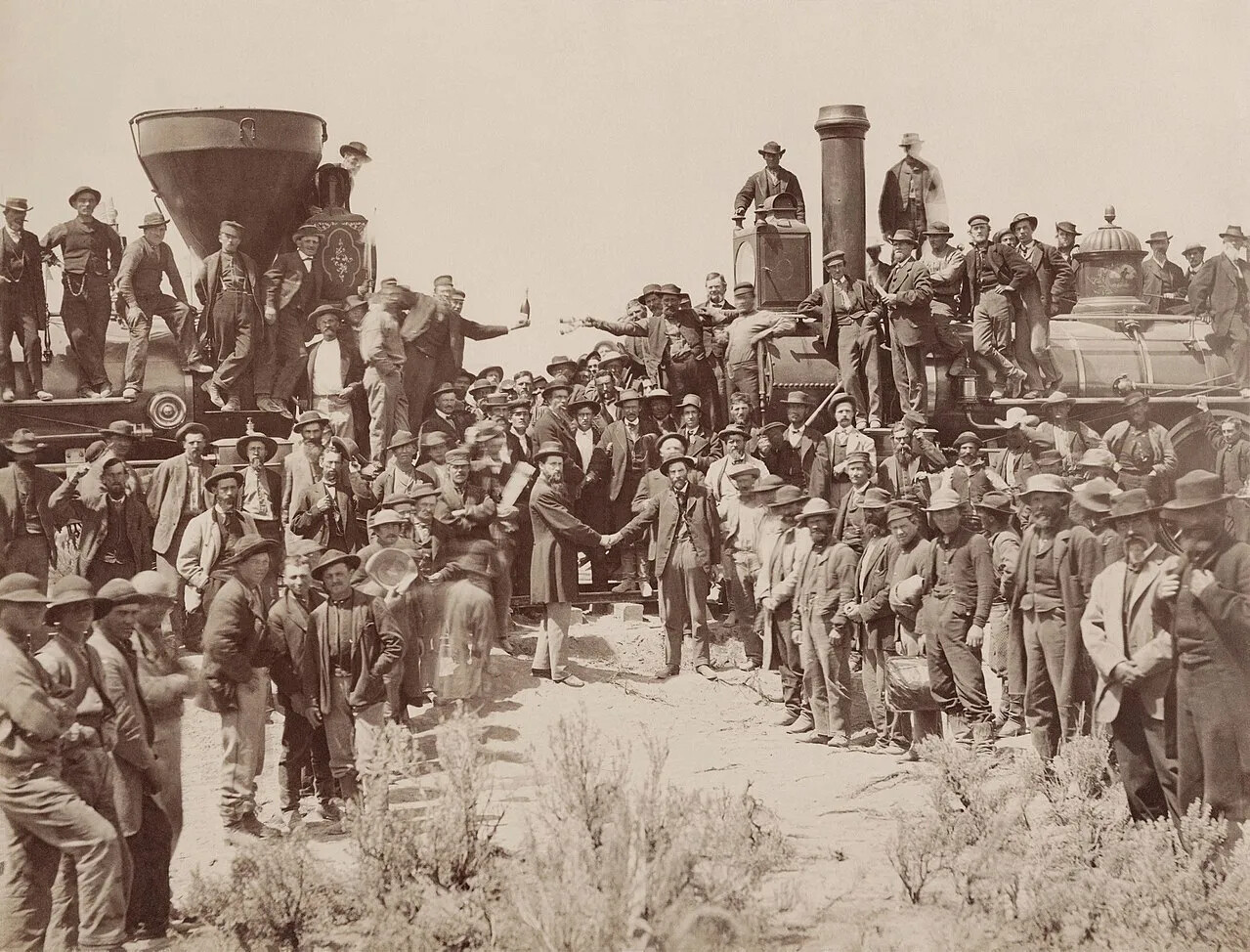 A black and white photograph shows two railroad engines facing each other. The left exhaust pipe is large and shaped like an inverted funnel, while the other exhaust pipe is straight. There are many men in the photograph, standing on the ground, on the engines, and all around. They generally wear hats and working clothes, and some carry shovels or pickaxes. Some are dressed as conductors on trains. Two men in suits stand in the center and shake hands while everyone watches them or faces the camera.   