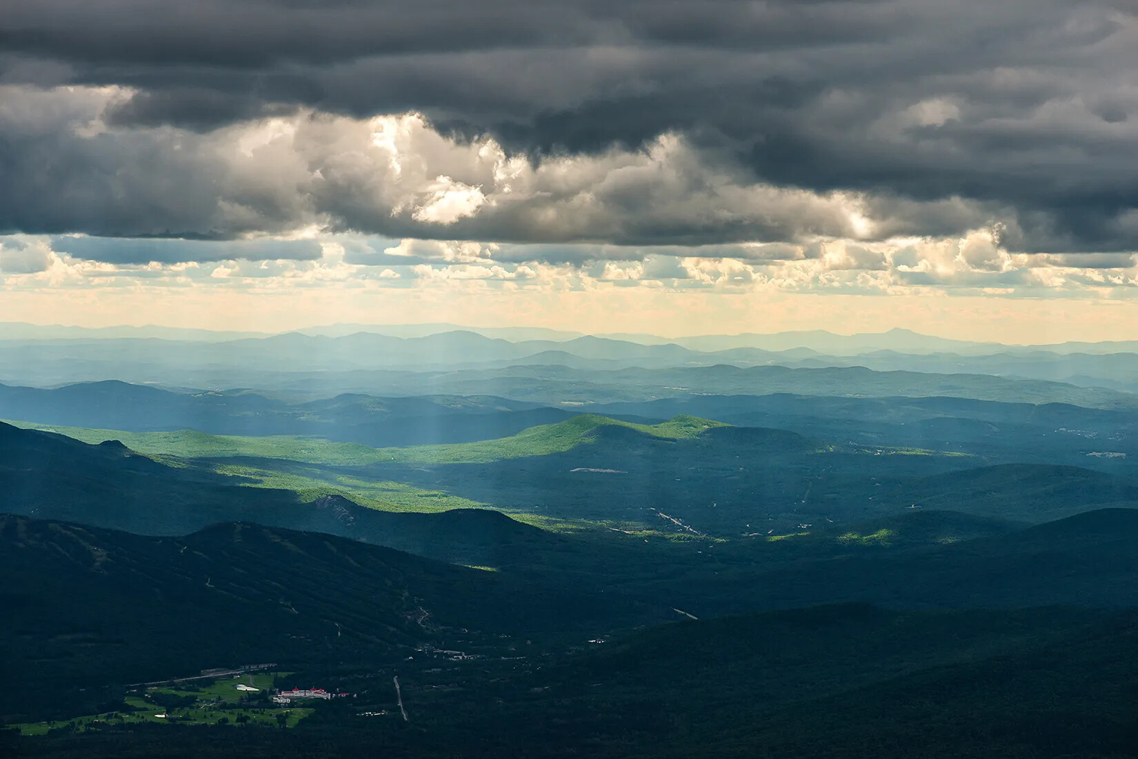 A colored photograph of a mountain range with dark gray clouds covering the upper border. Pockets of sunlight shine through the clouds down onto green spaces on the mountains.