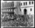 Part of a large stately building with columns and multiple entrances is seen in a black and white photograph. A large crowd of people have gathered on the street, and most wear wide brimmed hats, suits, and long jackets. Three 1930s cars are among the crowd. 
