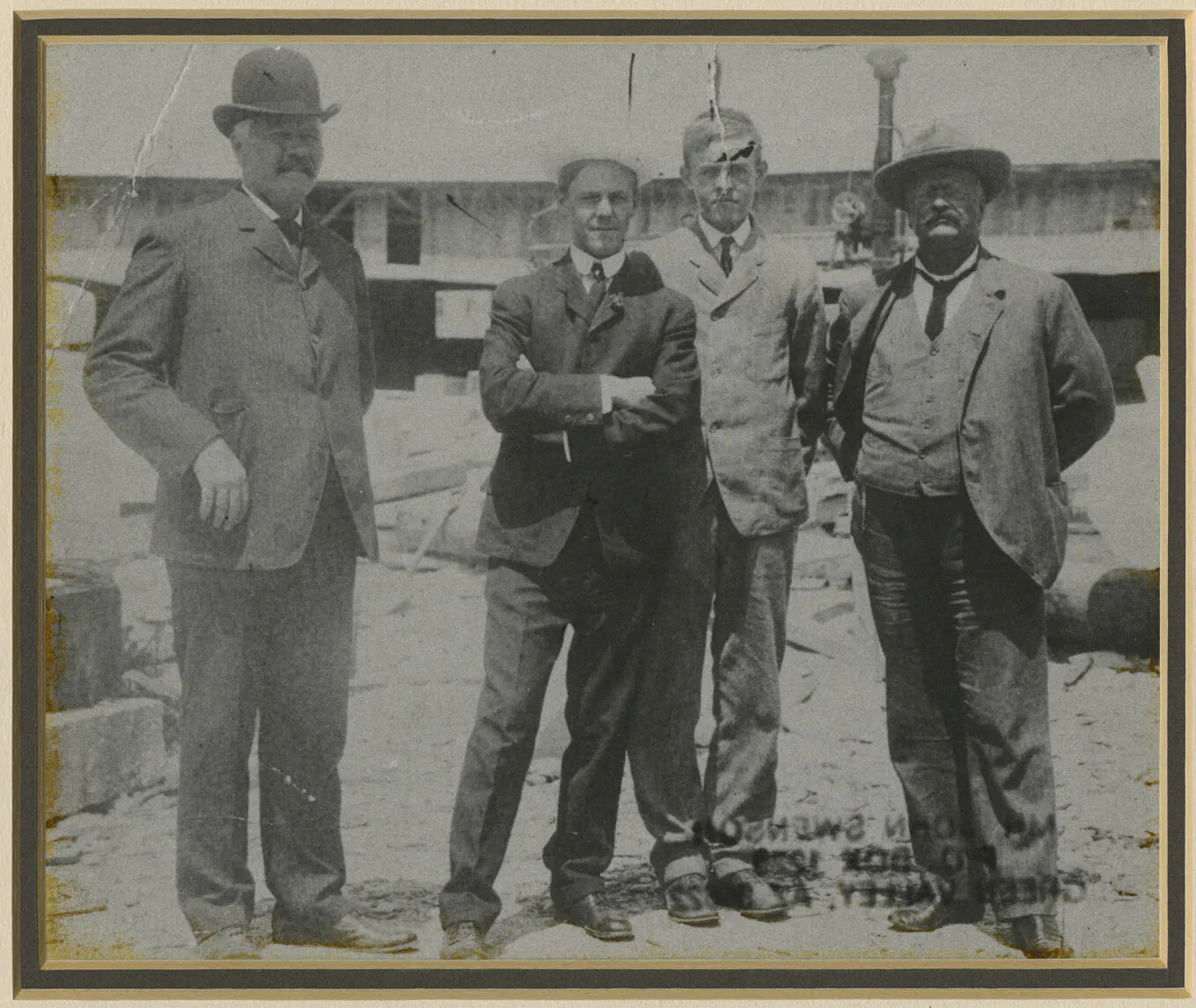 Four men in suits stand looking at the camera. The two men in the middle are younger, and stand together. The men generally have their hands behind them, and they are looking at the camera. The backdrop has gravel,  granite slabs, and a building.
