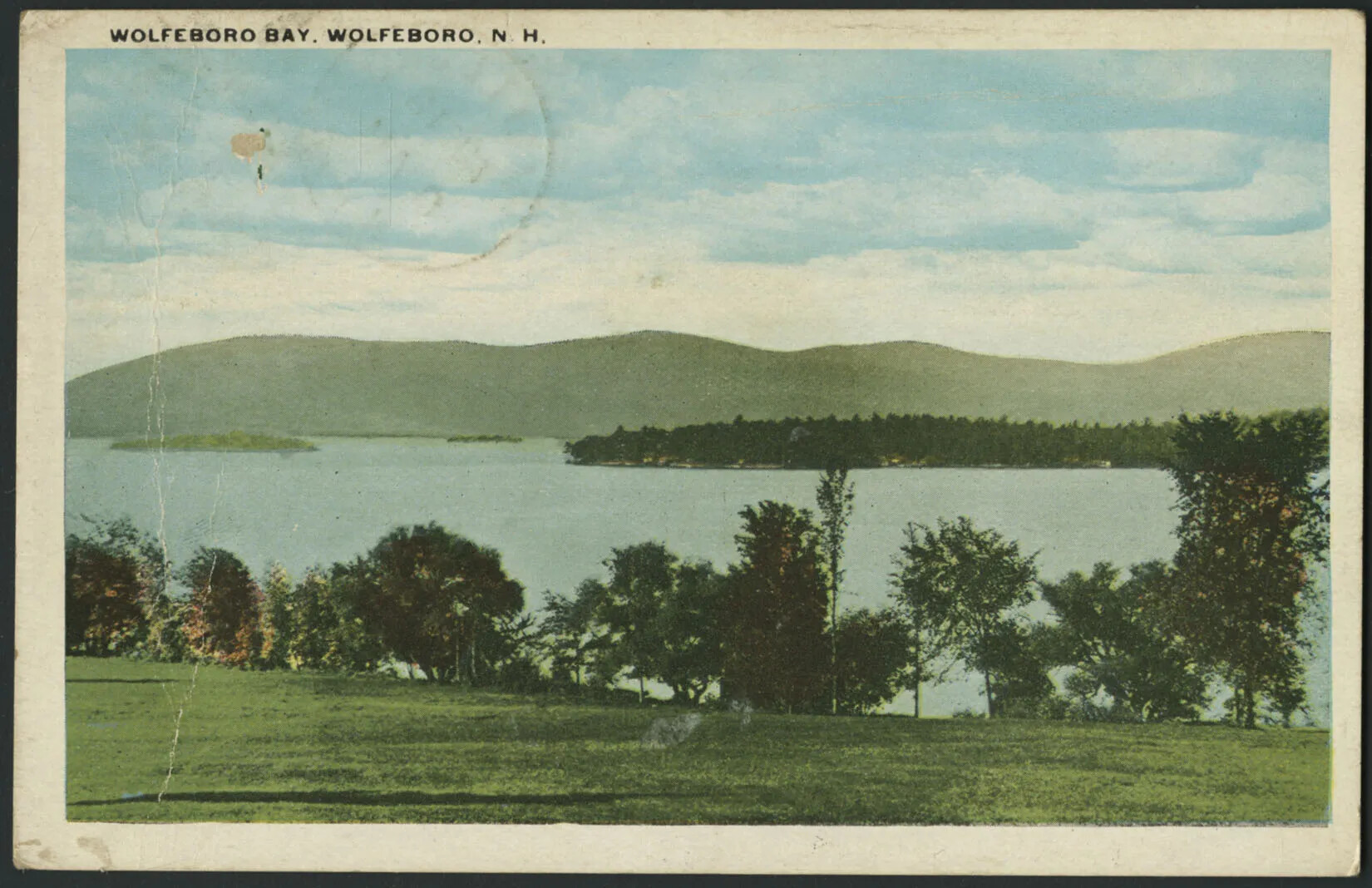 A colored postcard of a body of water surrounded by open green space, trees, and mountains. In the water are two green islands. The words "WOLFEBORO BAY. WOLFEBOROR, N. H." are written in black text in the top left-hand corner.