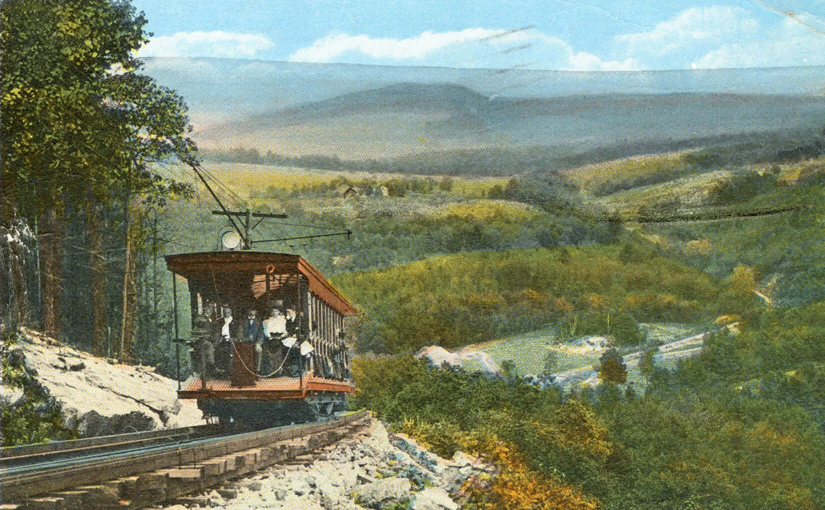 A colored horizontal postcard of a trolley filled with people, traveling up a steep mountain side on a set of railroad tracks. In the background, there is a deep valley filled with green trees and a large mountain in the distance. Above the mountain is a blue sky filled with white clouds.