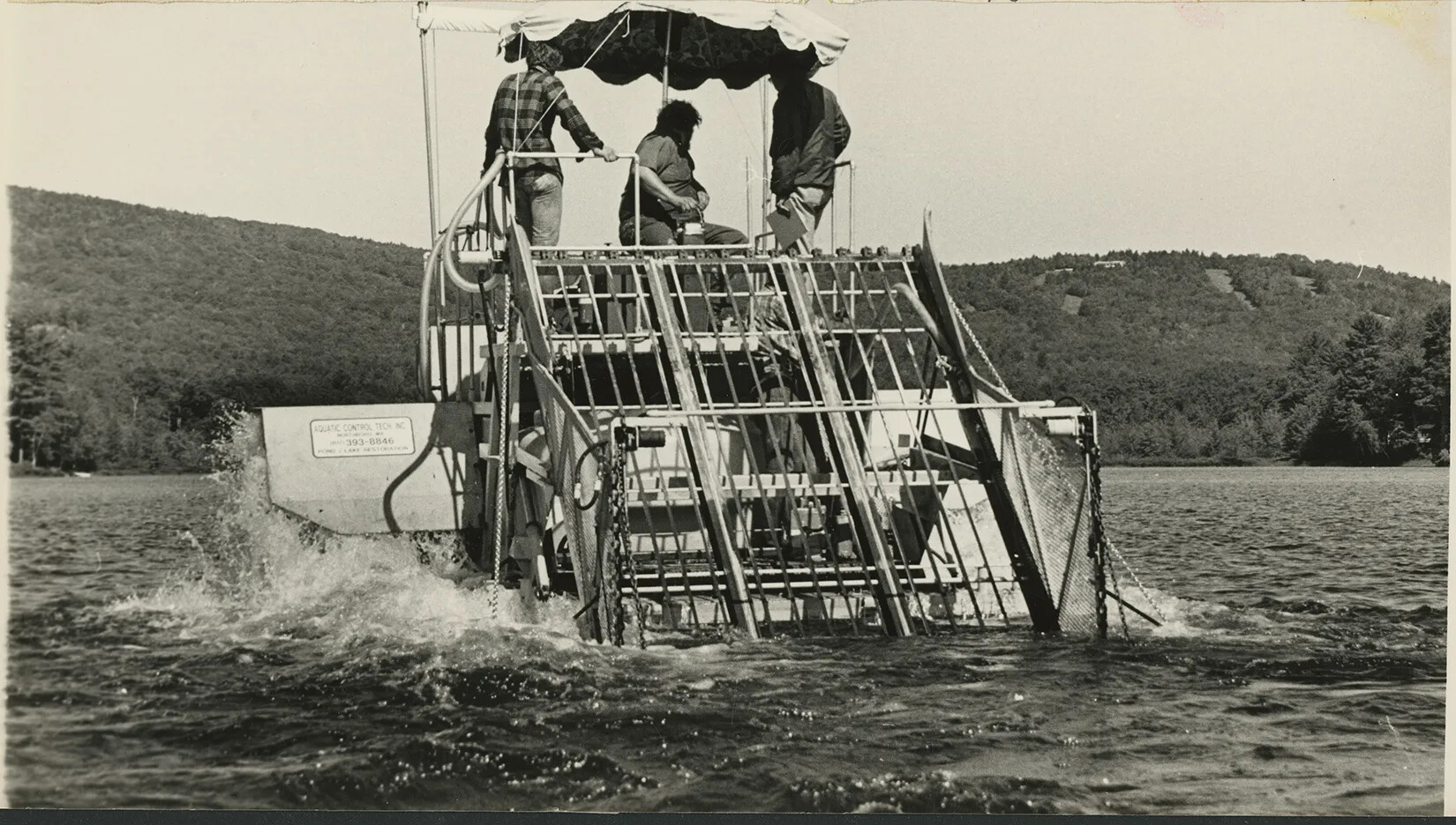 A black and white photograph of three people riding on a boat in a body of water surrounded by mountains. The boat has a large, cage-like contraption at the back of it.