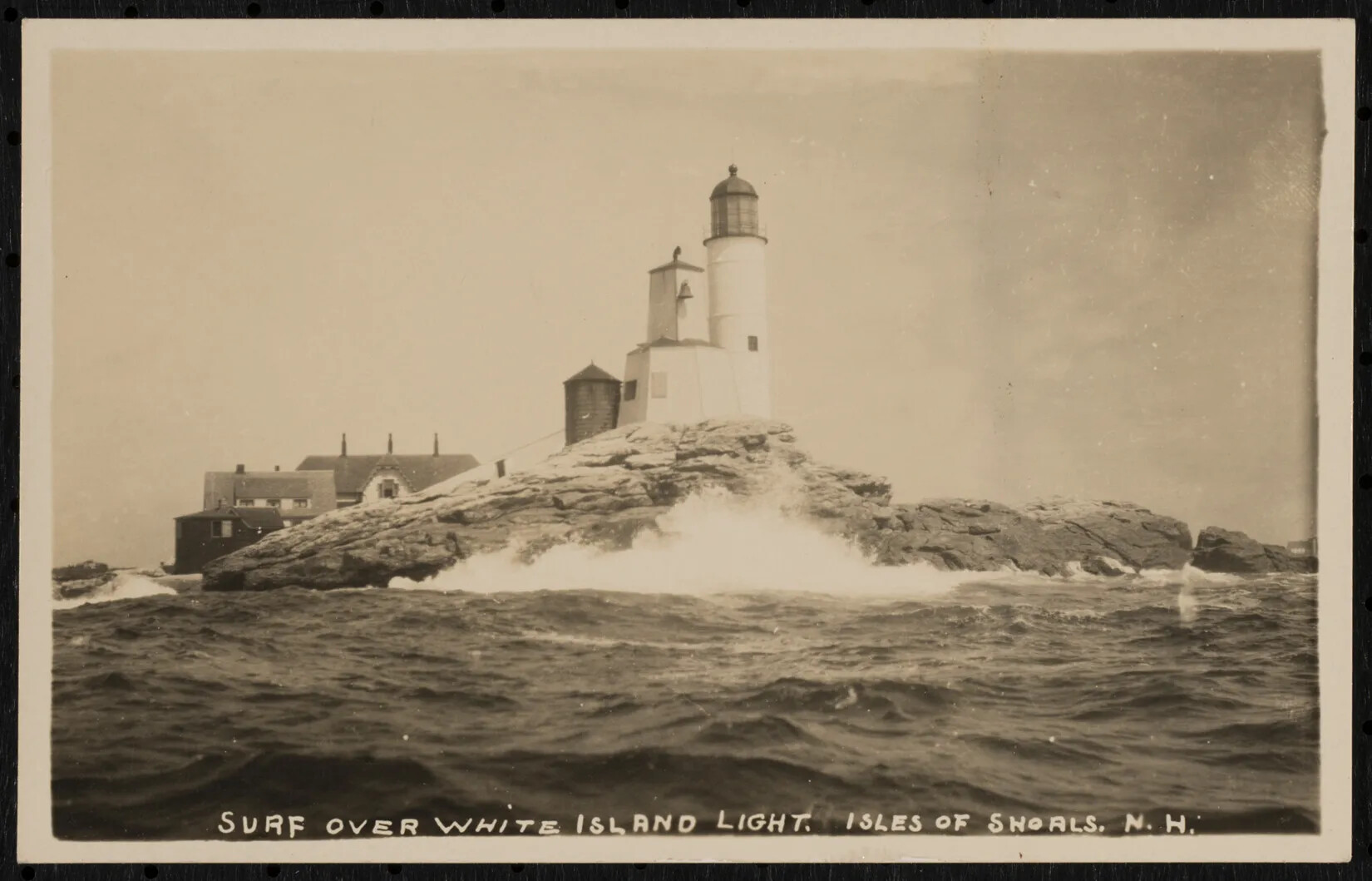A black and white photograph of a light house sitting atop a mound of rocks. The rock mound is surrounded by a body of water. Behind the lighthouse, to the left, are three small buildings. At the center, waves crash onto the rocks in front of the lighthouse. The photograph is framed on white paper. Words written in white along the base of the photograph read "SURF OVER WHITE ISLAND LIGHT. ISLES OF SHOALS, N.H."