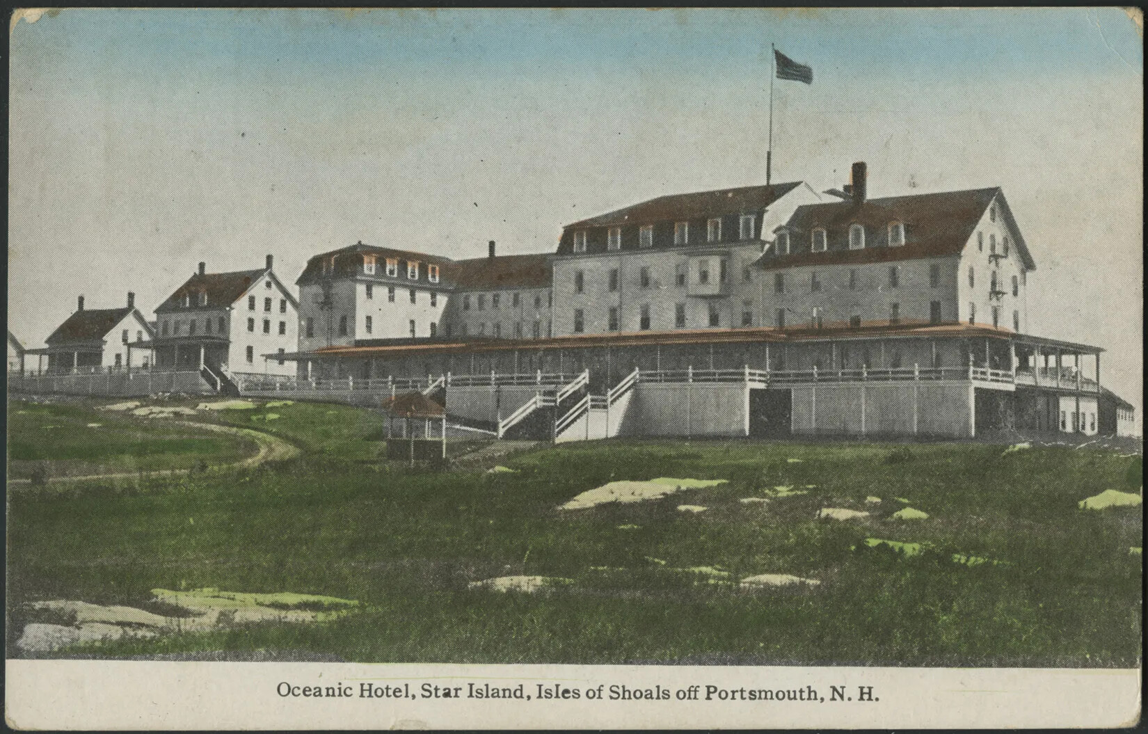 A color postcard of several attached and free-standing white buildings of varying sizes and red roofs. A tall flag pole and American flag flies over the building, and a large raised and covered porch circles the four right-most buildings. The two stand-alone buildings each have raised and covered porches. The foreground of the postcard shows a curved road in the left-hand corner and white rocks scattered in the grass. The painting is mounted on white paper. Words along the bottom edge of the postcard read "Oceanic Hotel, Star Island, Isles of Shoals off Portsmouth, N.H."