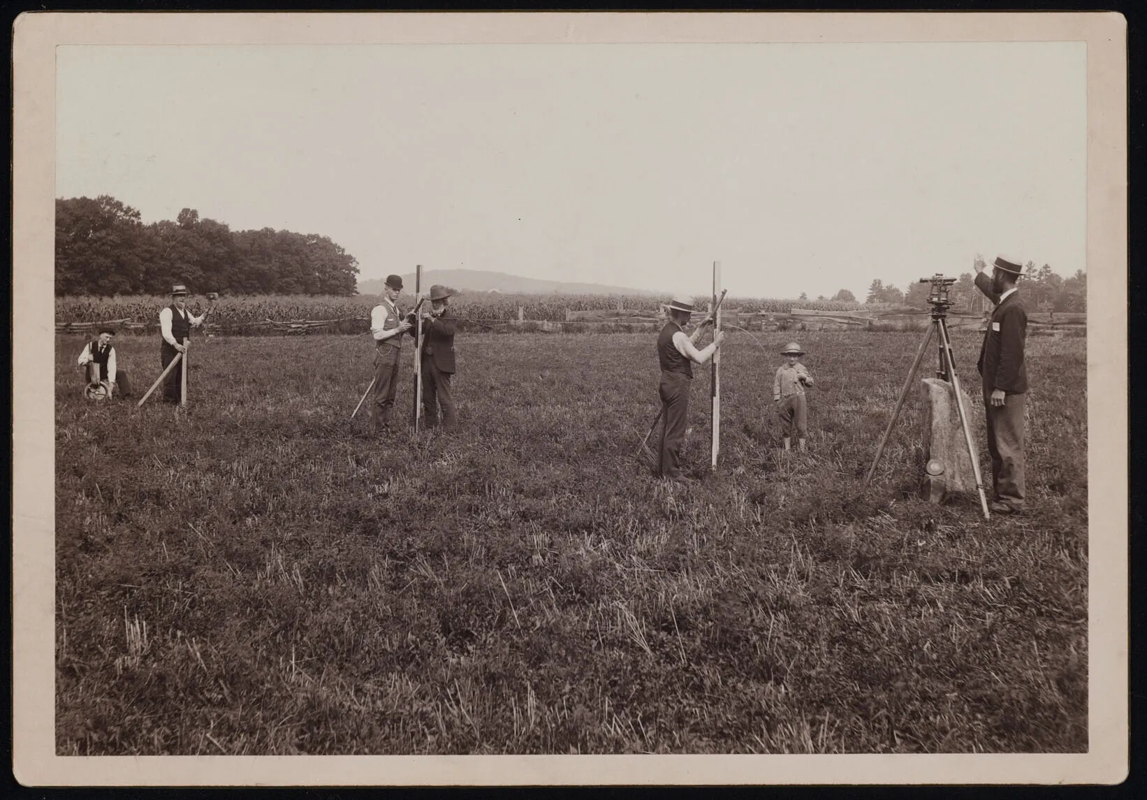 A black and white photograph of six men and one child, who are all using wooden tripods in a large, flat grassy field. Two tripods in the left-hand side of the photograph have two men standing at it, and two tripods on the right-hand side have one person standing at them. The child stands between the two men on the right side of the photograph.
