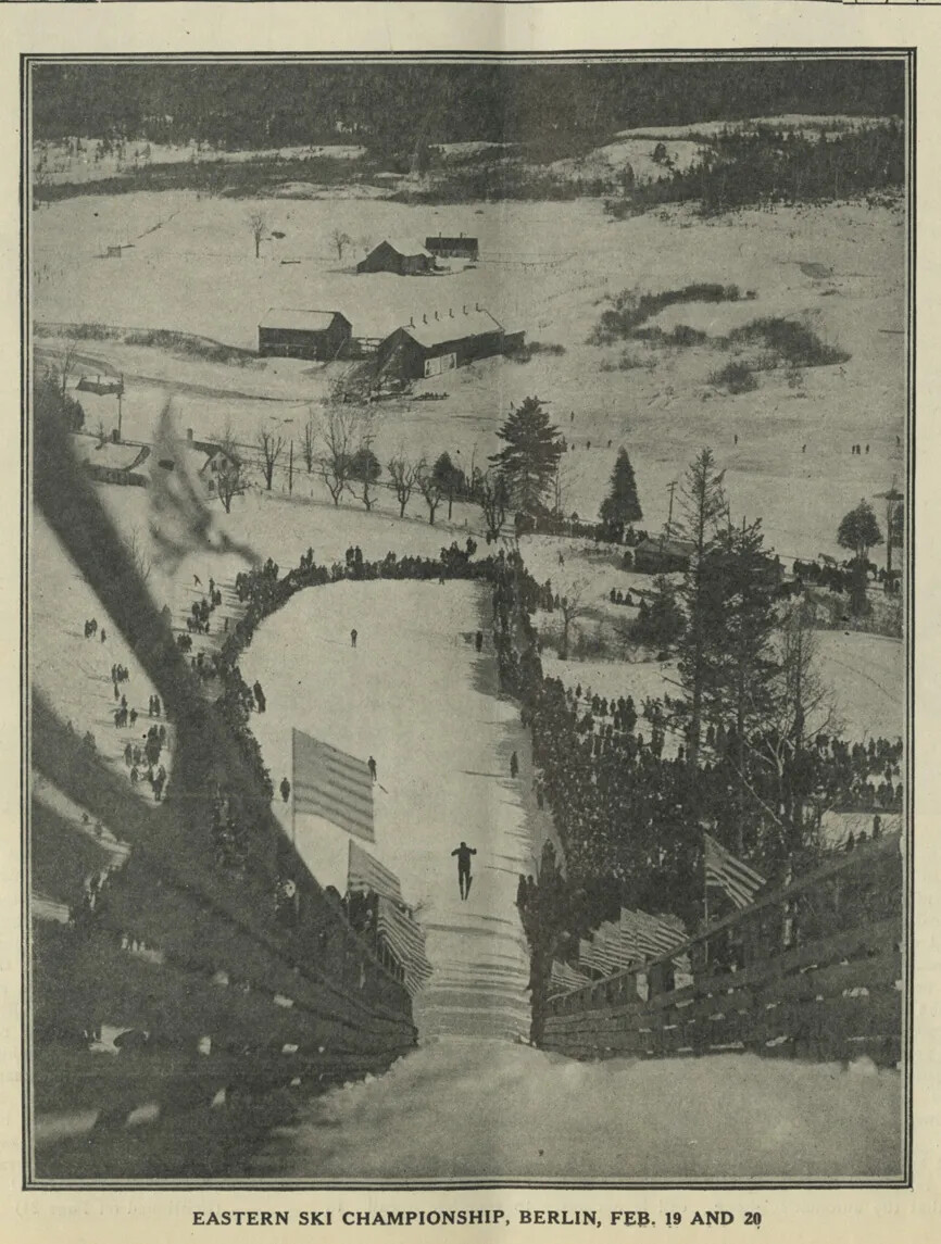 A black and white image of a skiing competition is shown. The photograph was taken above the spectators and skiers, on top of a hill.  The crowds of people are gathered around the ski slope, and a single skier is seen in the middle of the photograph.  The ski slope is sectioned off by wooden fencing and several American flags are posted along the fencing.  In the background, there are some farm buildings and forest in the distance.  Beneath the photograph is the caption: “EASTERN SKI CHAMPIONSHIP, BERLIN, FEB. 19 AND 20”.