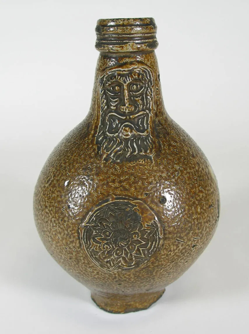 A brown shiny jug with the image of an old man's head carved into the neck of the jug. A circular geometric design is carved into the center of the jug.