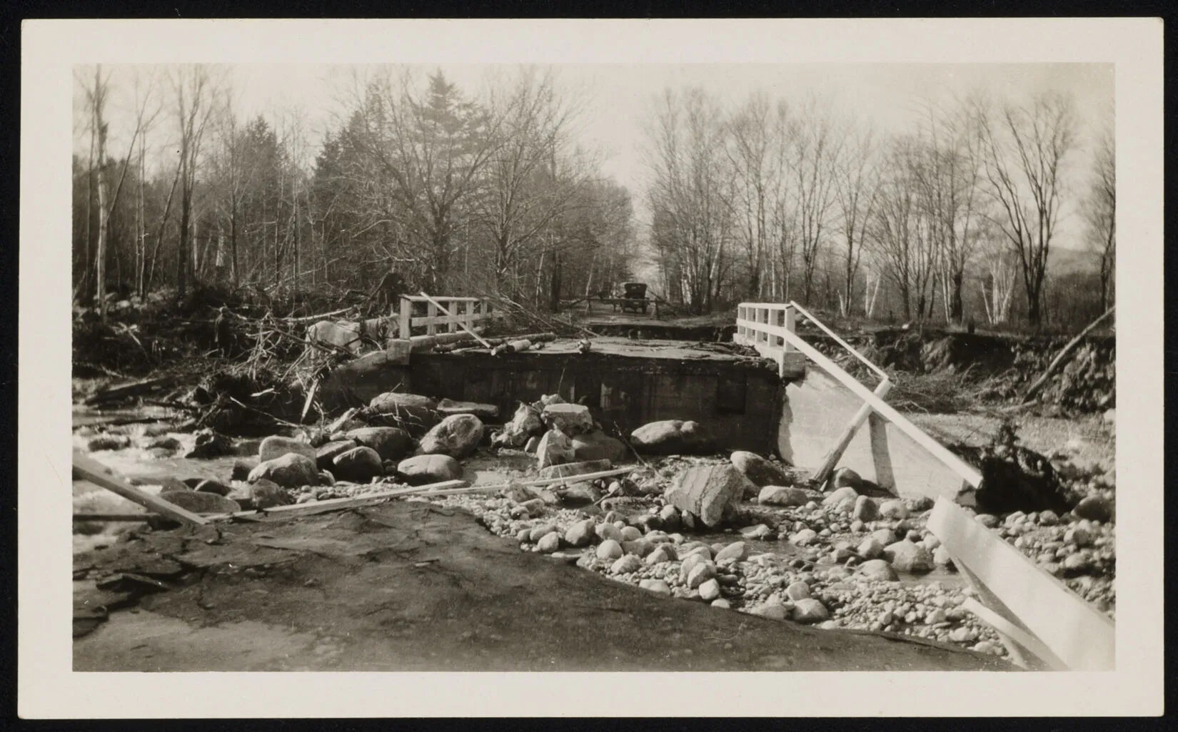 A black and white photograph of a collapsed bridge. The middle of the bridge is filled with large rocks. A truck is seen in the background stopped at a barrier.
