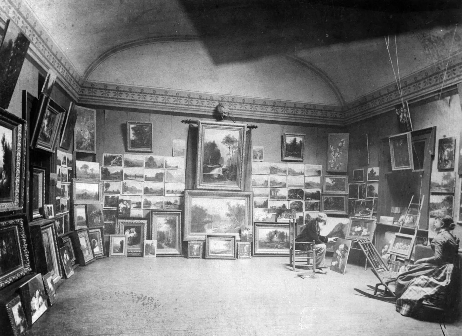 A black and white photograph of the interior of an art gallery. The walls are covered in paintings of many shapes and sizes. A man and a woman sit at a distance from each other along the right side of the photograph, each looking at a painting placed in front of them.