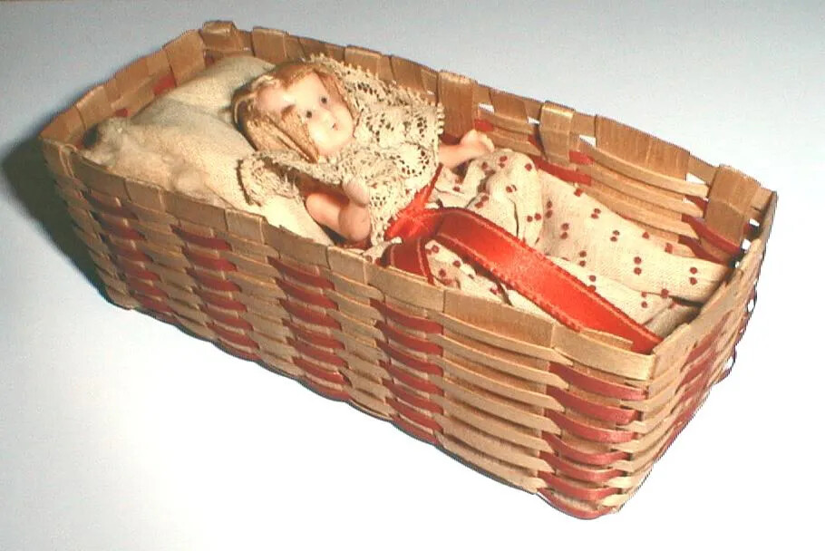 A woven basket with a baby doll laying down on a plush, white blanket. The woven pattern on the basket alternates thin strips of red and natural colored pieces of wood. The baby doll in the basket wears a white dress with red polka dots and a large red sash tied around the middle.