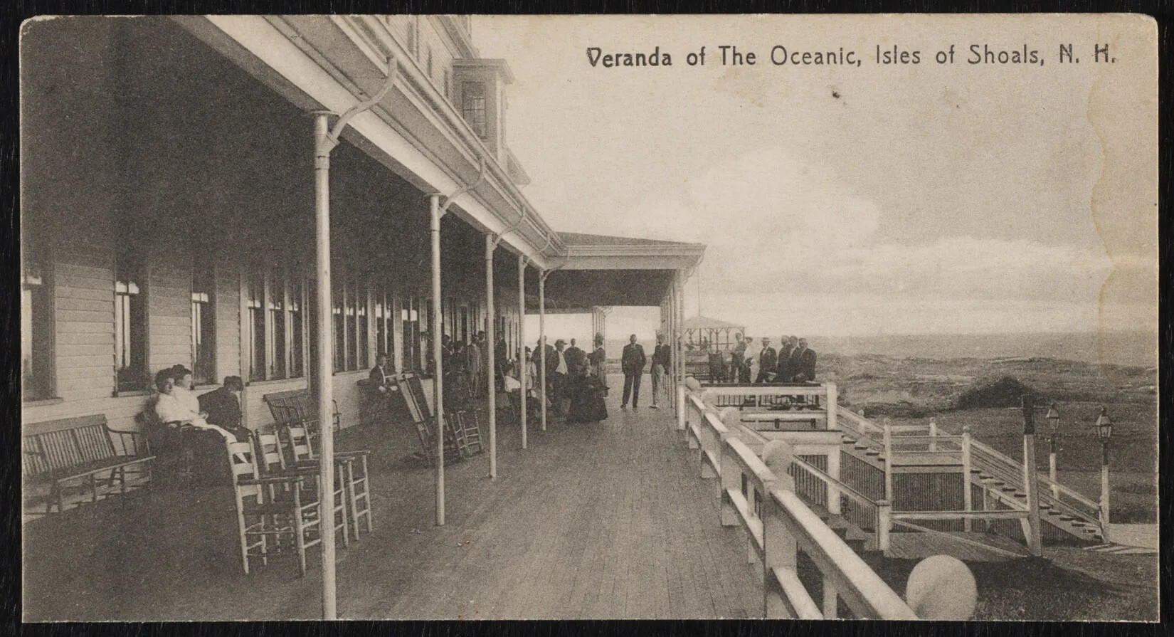 A black and white photograph of a large front porch. A number of people are sitting and standing around the porch, and there are several empty benches and chairs placed underneath the covered part of the porch. A set of stairs leads down to a path and grassy area in the right-hand corner. Two tall lanterns stand at the end of the stairs at ground level. The title of the photograph, which appears in the top right corner, reads “Veranda of The Oceanic, Isles of Shoals, N. H”.