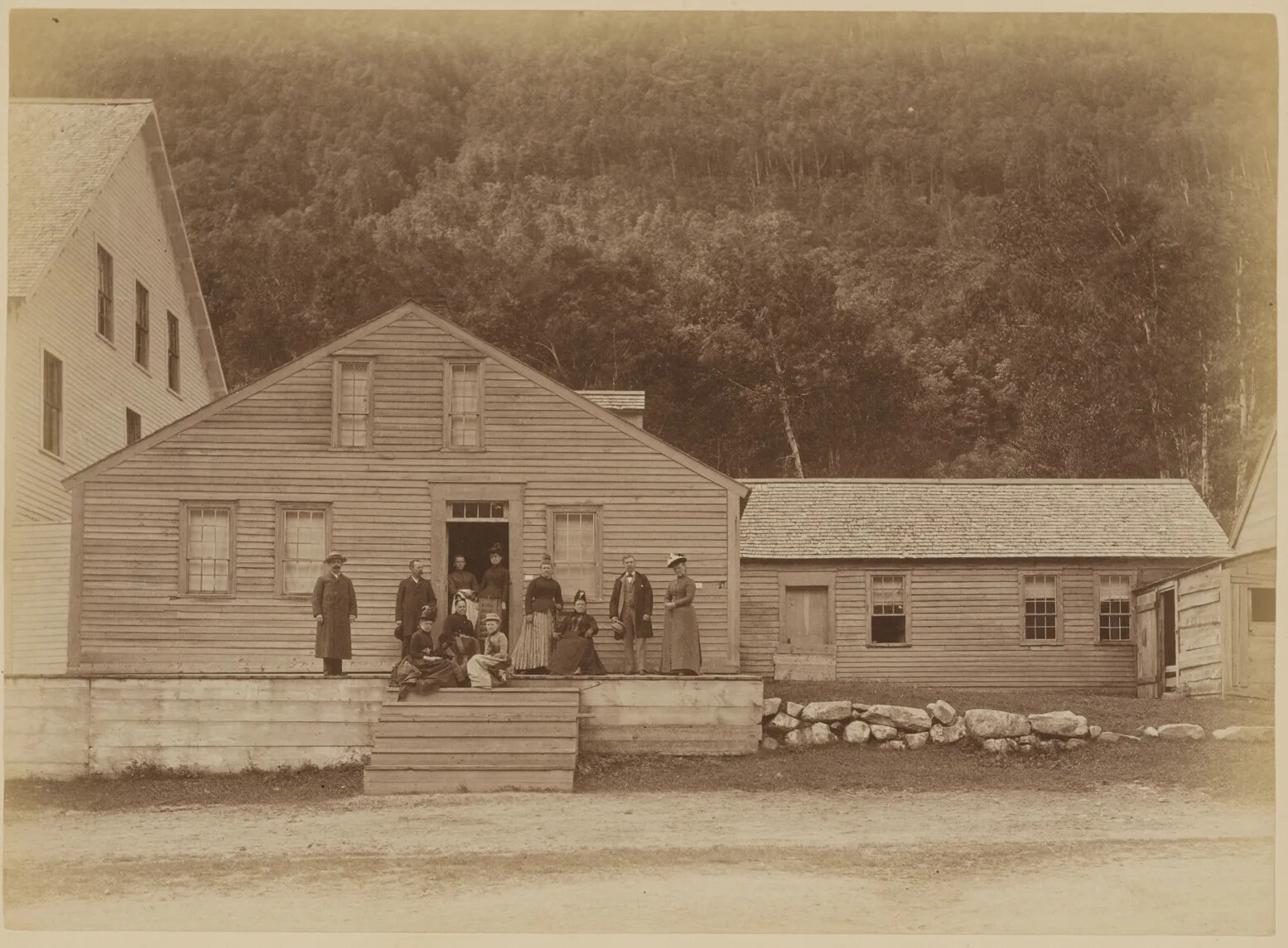 Black and white photograph of a group of people in front of a small, two-storied building with a pointed roof. Some people stand on the raised platform in front of the house, while others sit in either a chair or on the steps leading up to the house from the ground-level. To the left of the house is a larger building, and to the right is a low stone wall along the grass. Behind and attached to the smaller building is a one-story "L" shaped building and shed. Behind the buildings is forested area. The photograph is mounted on plain white paper.