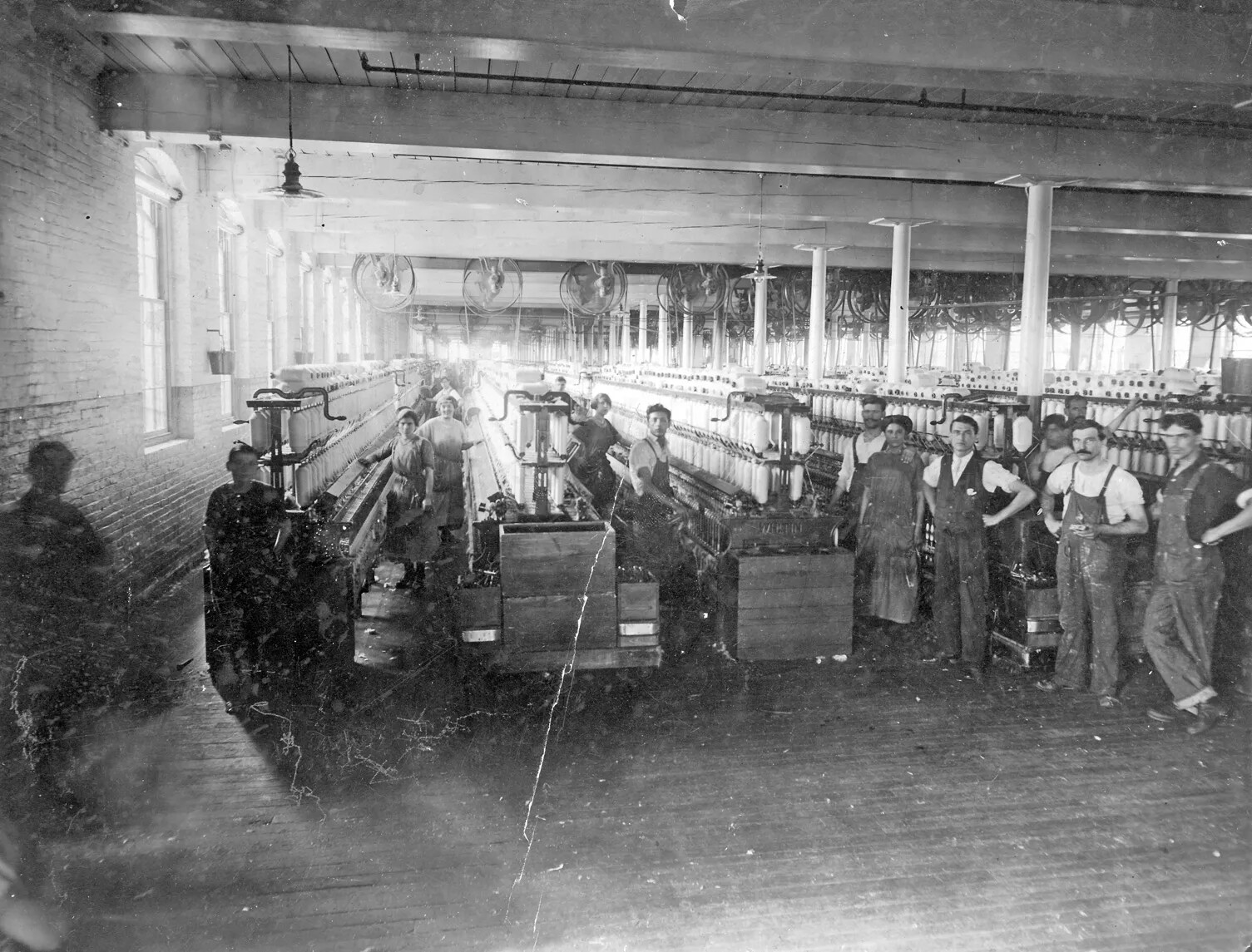 A black and white photograph of a group of white men and women inside a textile factory. There are columns of machinery with cylinders of white yarn at the tops of the machines. The women all wear long dresses and the men wear short-sleeved shirts and overalls. One man in the right side of the photograph is wearing a collared shirt with a vest.