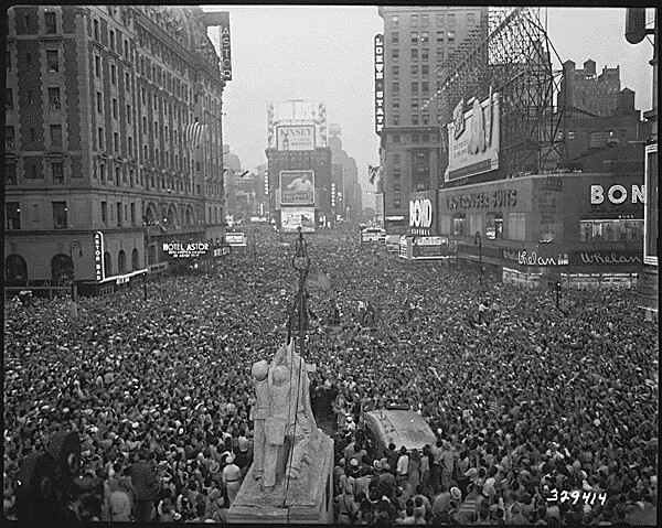A black and white photograph of a wide street filled with people. The street is lined with multi-storied buildings.