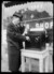 A black and white photograph shows a man in a long coat and hat standing outside a store on a street. Next to him is a table with a large black box on it. On the box are two circles with markings. He is holding something connected with a wire to the black box up to his ear, and touching one of the circles with the other hand. He is looking down at the box.. 