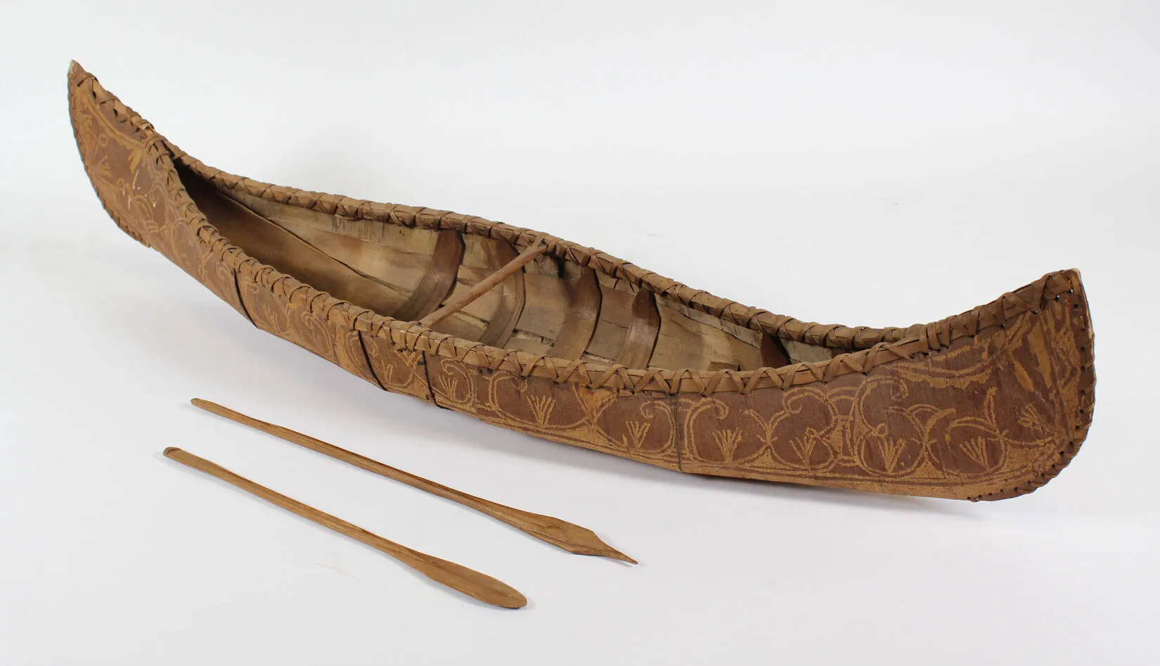 A small wooden canoe with ornate carvings on the exterior. A thin wooden bar lies across the inside of the canoe, in the center. Two small wooden oars lie to the left of the canoe.