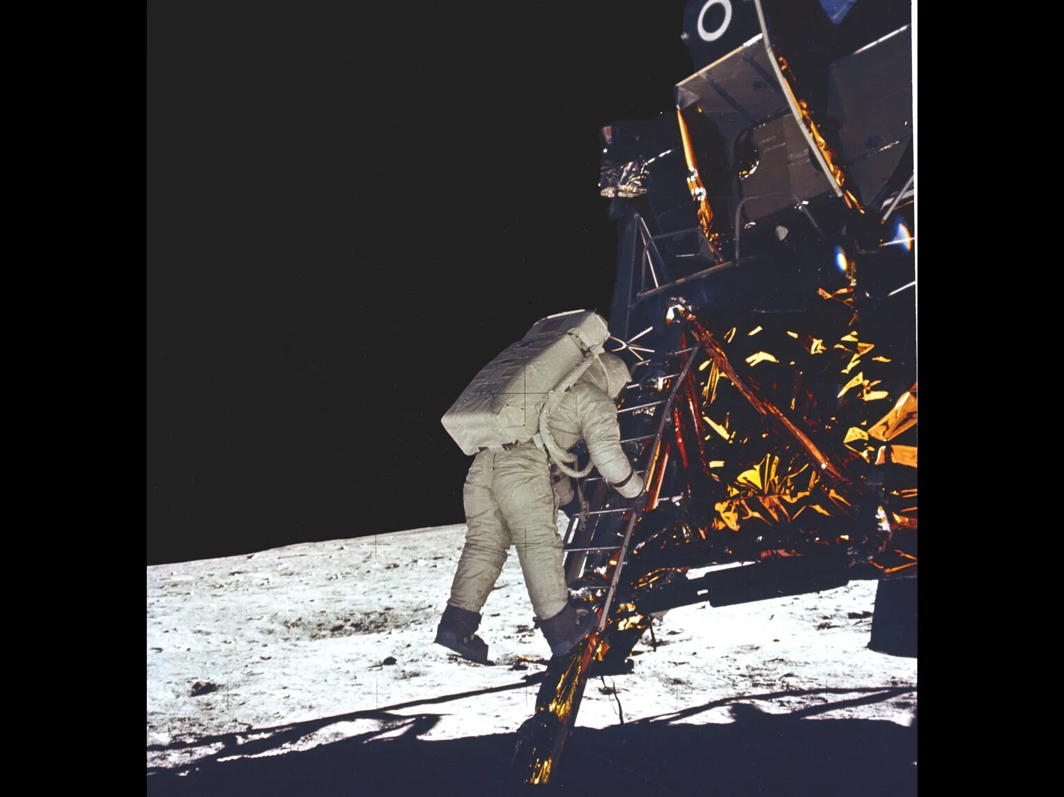 In a color photograph, an astronaut with a white spacesuit and large backpack descends a ladder. He is leaving a small spaceship and stepping down to walk on the moon. 