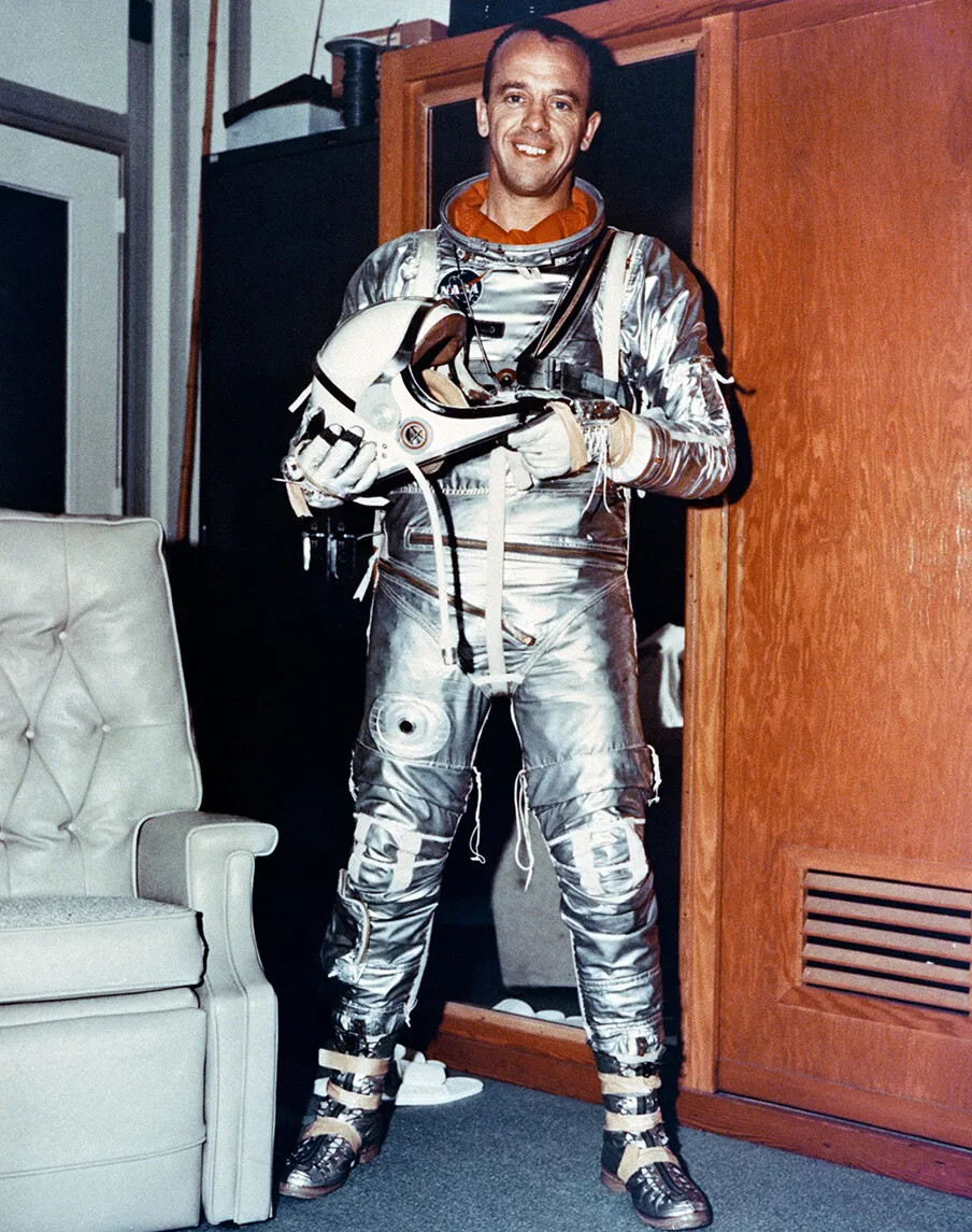 A colored photograph of a white man in an astronaut suit. He looks at the camera, and holds his helmet in his hands.