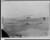 In a black and white photograph, a biplane is shown with two wings stacked on each other. A small engine is seen along with a small body of the plane behind the wings. The plane is on a beach and a man stands watching it, near a wooden bench. 