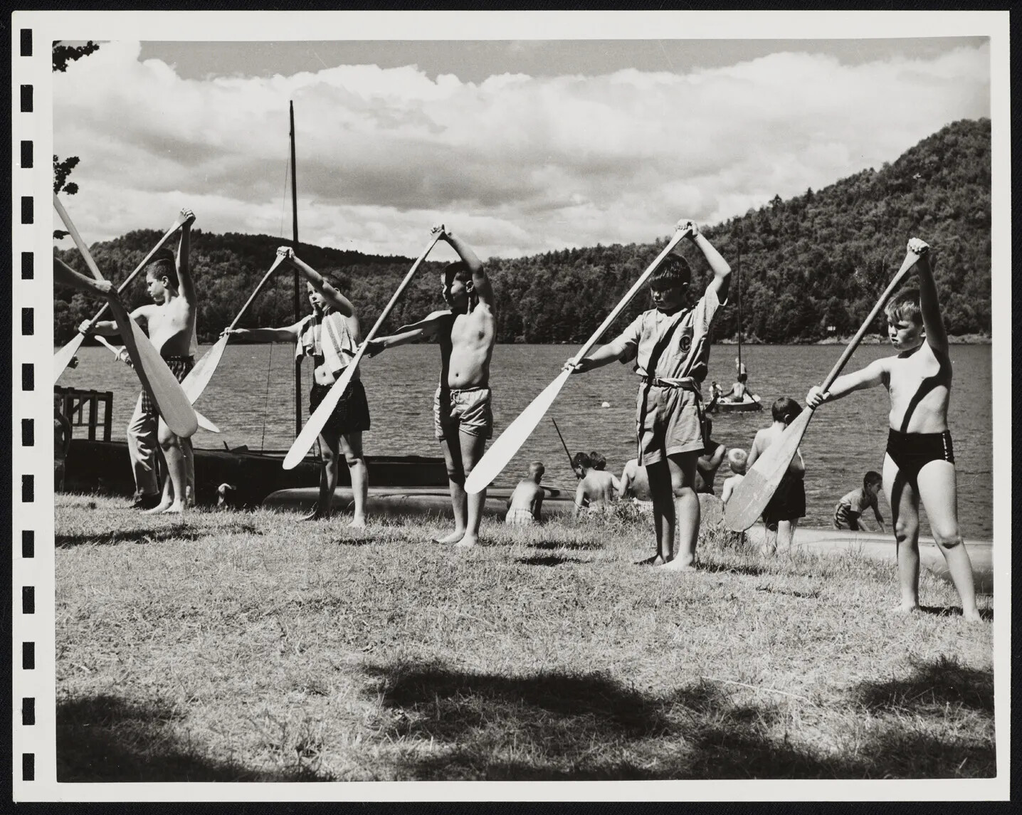 A black and white photograph of a group of children standing on a grassy area, holding large wooden paddles in their hands. In the background, there is a small body of water surrounded by mountains.