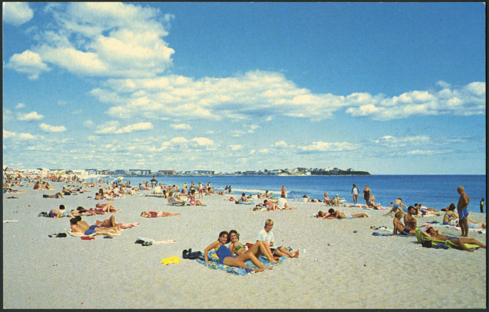 A colored horizontal postcard of a flat area of sand. Many people sit in groups on the sand. To the sand's right is a large, flat body of water. Along the back edge of the postcard are several buildings. A large blue sky with groupings of white clouds take up the majority of the top of the postcard.