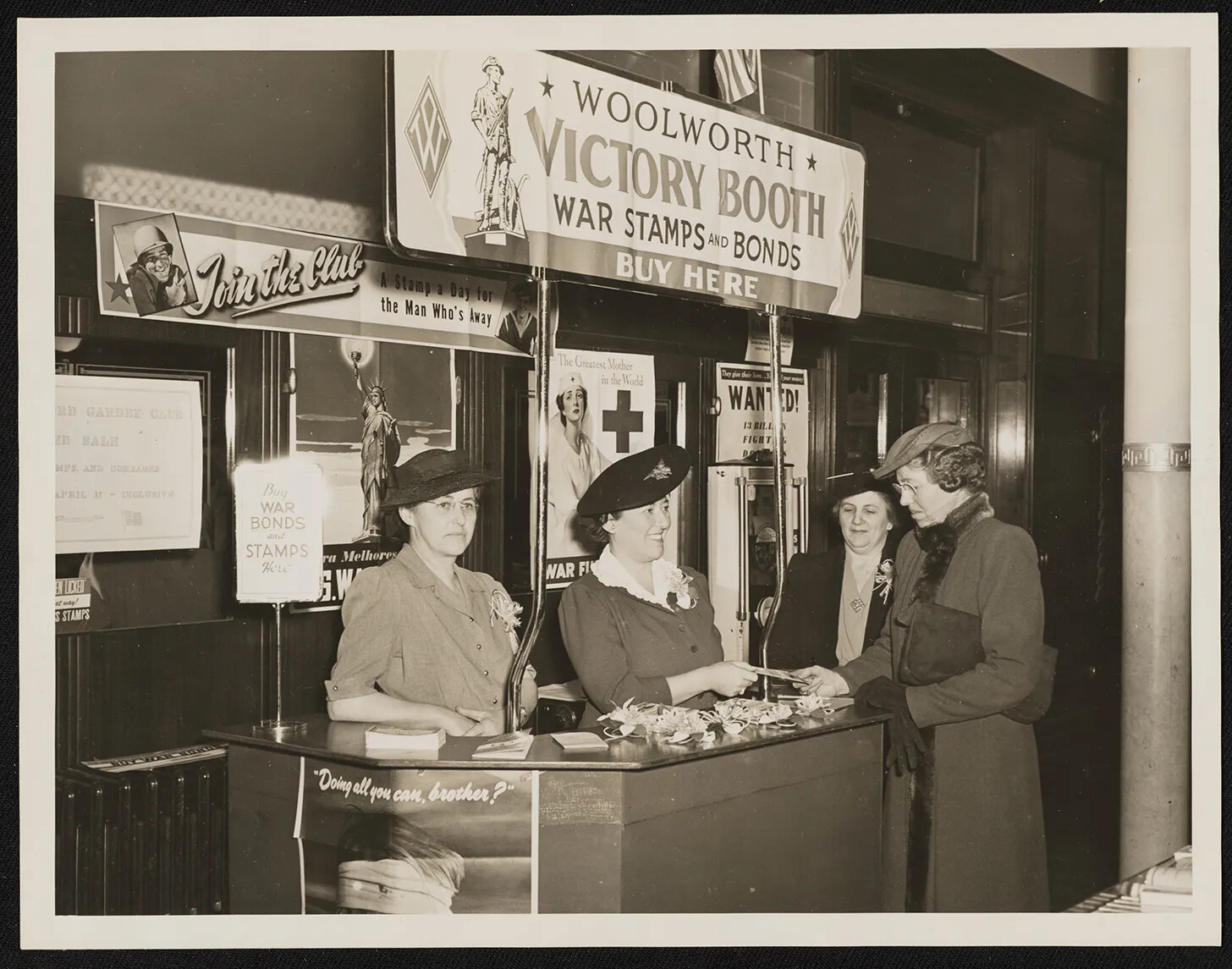 A black and white photograph of four women standing around a booth. A sign hanging above the booth reads "WOOLWORTH/VICTORY BOOTH/WAR STAMPS AND BONDS/BUY HERE."